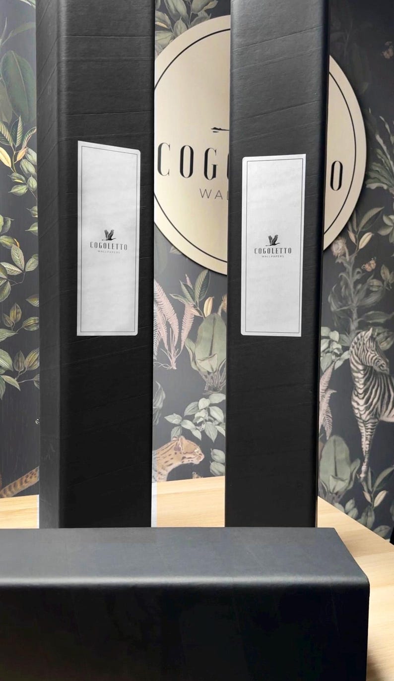 May include: Black rectangular boxes with white labels featuring the "COGOLLETO WALLPAPER" logo. The background showcases a wallpaper design with a jungle theme, including plants and animal illustrations. The boxes are on a light wood surface.