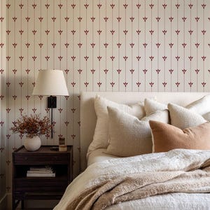May include: A bedroom scene featuring a bed with a cream-colored headboard and bedding. The bed is adorned with several pillows in varying shades of cream and tan. A dark wood nightstand holds a lamp and decorative items. The wallpaper has a cream background with a repeating burgundy floral pattern.