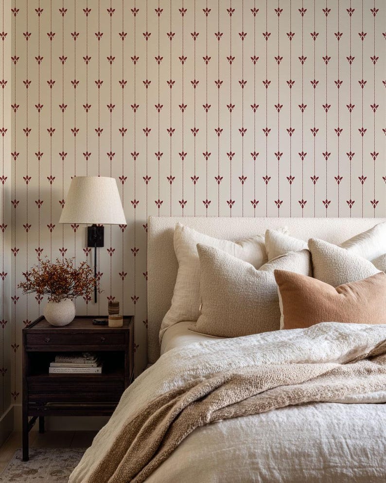 May include: A bedroom scene featuring a bed with a cream-colored headboard and bedding. The bed is adorned with several pillows in varying shades of cream and tan. A dark wood nightstand holds a lamp and decorative items. The wallpaper has a cream background with a repeating burgundy floral pattern.