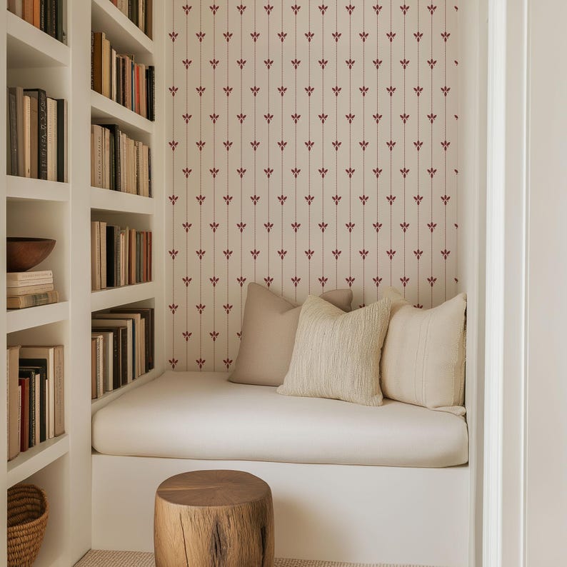 May include: A cozy reading nook with built-in white bookshelves filled with books. A white cushioned bench is adorned with beige pillows, set against a patterned wallpaper. A wooden stool sits in front of the bench.