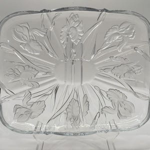 May include: Clear glass serving dish with a rectangular shape and a floral iris design. The dish has a raised, frosted iris pattern and is divided into two sections. The edges of the dish have a scalloped design.