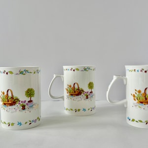May include: Three white ceramic mugs featuring a repeating pattern of a brown wicker basket filled with orange and yellow flowers, a small potted green tree, and purple flowers. The mugs have a decorative border of green and blue leaves.