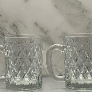 May include: Two clear glass mugs with diamond-shaped cut patterns. Each mug has a large, curved handle. The mugs are displayed against a white and gray marble-like background. These mugs are suitable for beverages.