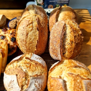 May include: A close-up of six loaves of freshly baked sourdough bread. The loaves are golden brown and have a rustic appearance. Some of the loaves have a decorative pattern on the top.