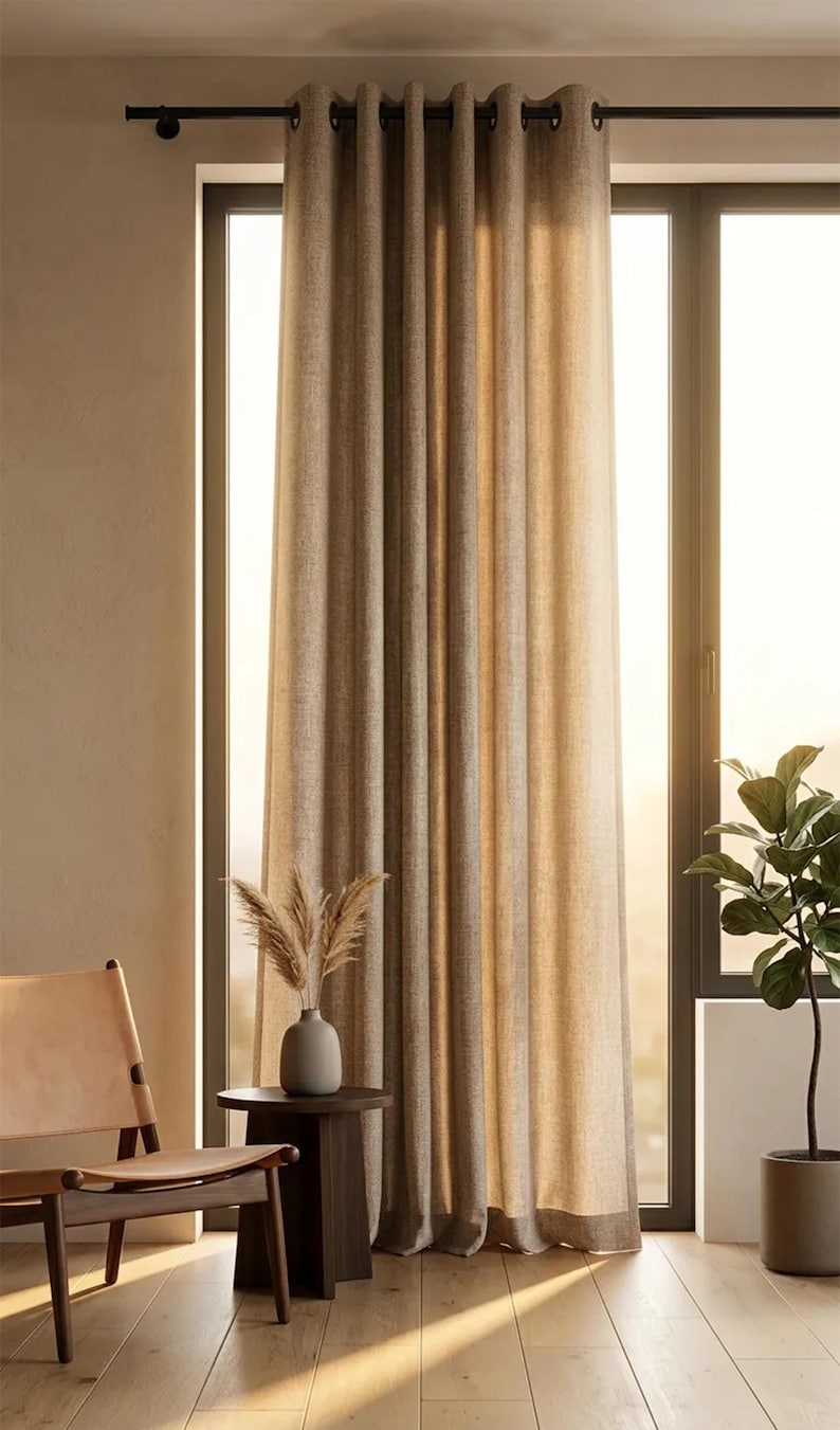 K&ouml;nnte beinhalten: Beige Leinen-Vorh&auml;nge mit &Ouml;sen h&auml;ngen vor einem gro&szlig;en Fenster an einer schwarzen Stange. Ein kleiner Beistelltisch h&auml;lt eine Vase mit getrockneten Pflanzen. Ein Lederstuhl und eine Topfpflanze befinden sich ebenfalls im Raum.