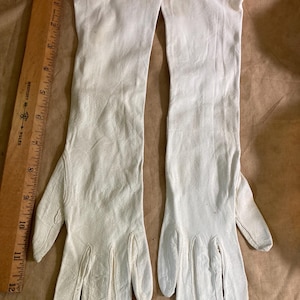 May include: A pair of long, white opera-length gloves with a smooth texture and stitched finger details. A wooden Westcott ruler is positioned alongside the gloves, showing a length of approximately 12 inches for the glove portion.