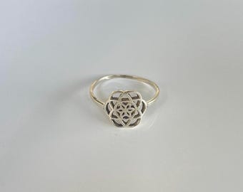 Op de afbeelding: Een zilveren ring met een bloemmotief. De ring heeft een smalle band en een gedetailleerde, opengewerkte bloem in het midden. Het bloemontwerp is symmetrisch, met meerdere lagen bloemblaadjes. De ring staat tegen een witte achtergrond.