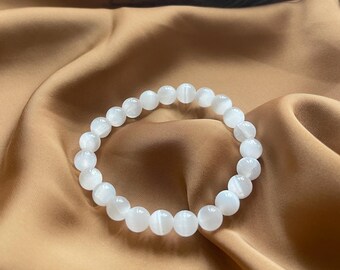May include: A white gemstone bracelet with round beads. The bracelet is on a brown fabric background.