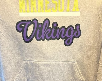 May include: A grey hoodie with the words "MINNESOTA" in yellow block letters above "Vikings" in purple script with a black outline. The hoodie has a front pocket.