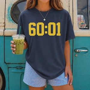 May include: A dark charcoal gray t-shirt featuring the yellow text 60:01 in a distressed block font. The shirt is paired with blue and white vertical striped shorts. A clear plastic cup filled with a green iced beverage is held in one hand.