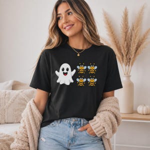 May include: A black short-sleeved t-shirt featuring a graphic print of a white cartoon ghost on the left side and four small yellow and black striped bees with blue wings arranged in a square pattern on the right side of the chest area.