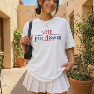 Op de afbeelding: Een wit T-shirt met korte mouwen en de tekst VOTE PAUL 4 BAKER in het rood en blauw op de borst gedrukt. Het shirt wordt gecombineerd met een witte geplooide rok en een zwarte schoudertas voor een eenvoudige en strakke uitstraling.