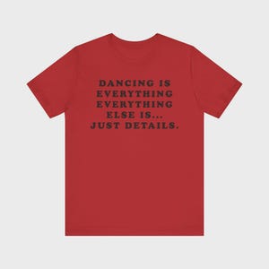 May include: A red short-sleeved t-shirt featuring black text centered on the chest that reads: DANCING IS EVERYTHING EVERYTHING ELSE IS... JUST DETAILS. The garment has a crew neckline and a standard fit, showcasing a bold contrast between the red fabric and the dark lettering.