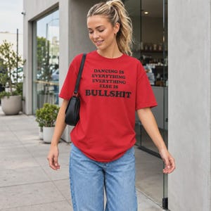 May include: A bright red short-sleeved t-shirt featuring the black printed text DANCING IS EVERYTHING EVERYTHING ELSE IS BULLSHIT on the front, paired with light blue denim jeans and a black shoulder bag with a thin strap.