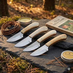 May include: Four hunting knives with bone handles and stainless steel blades are arranged on a dark stone surface. A rolled leather strap, a tin of beeswax, and a vintage field guide are also present, suggesting outdoor use.