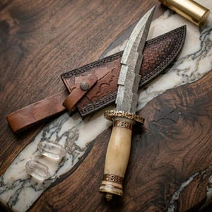 May include: A handcrafted dagger with a patterned steel blade and a bone handle, accented with gold-colored metal. The dagger rests on a wooden surface next to a brown leather sheath. Two clear quartz crystals are also visible.