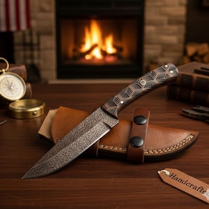 May include: A handcrafted knife with a textured blade and a patterned wooden handle, resting on a brown leather sheath. The knife features a dark, rustic finish. The sheath has two black snap closures. A tag that says "Handcrafted" is attached.