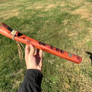 May include: A wooden flute with six holes and a leather strap. The flute is made of reddish-brown wood and has a natural finish.
