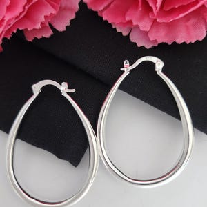May include: A pair of silver teardrop hoop earrings. The earrings are presented on a black cloth, with pink flowers in the background. The hoops have a smooth, polished finish and a classic design.