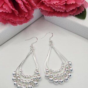 May include: Silver teardrop earrings with three tiers of small, round beads. The earrings have a simple, elegant design with a hook closure. The background features pink flowers.