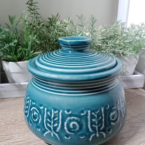 May include: Teal ceramic jar with a lid and a decorative pattern of white swirls and leaves. The jar is sitting on a wooden surface.