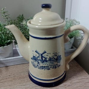 May include: A cream-coloured ceramic coffee pot with a blue and white windmill design. The pot has a blue lid and a blue handle.