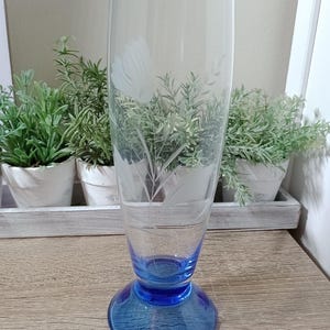May include: A tall, clear glass with an etched floral design and a blue base. The glass is ideal for serving drinks.