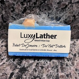 May include: A blue bar of Luxy Lather natural artisan soap, wrapped in clear plastic. The soap has a light blue hue with a white label that reads "Soleil De Janeiro / Do Not Disturb." The soap weighs 4 oz.