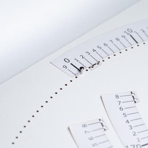 Könnte beinhalten: Eine weiße, kreisförmige Papierschablone mit einer gebogenen Skala von 0 bis 10, in die an der 0-Markierung eine kleine Metallnadel eingesteckt ist. Zwei weitere rechteckige Papierstreifen mit Zahlenskalen liegen daneben und zeigen Werte bis 230.