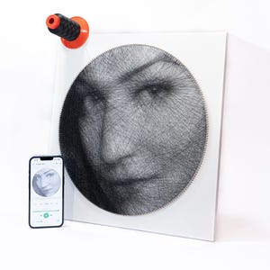Peut inclure: Un panneau carré blanc présentant un portrait en art filaire circulaire créé avec du fil noir enroulé autour de clous argentés. Une bobine de fil noire avec une base orange est placée en haut à gauche, et un smartphone affiche le portrait.