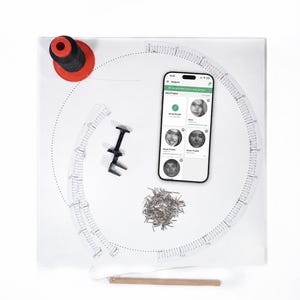 Puede incluir: Un tablero cuadrado blanco con una plantilla circular con marcas numeradas, un carrete de hilo negro sobre una base roja, un teléfono inteligente con una aplicación de arte con hilos, una herramienta de metal negro, clavos plateados y un palo de madera.