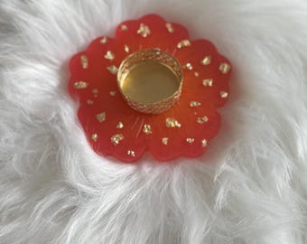 May include: A flower-shaped resin coaster in shades of red and orange, decorated with gold leaf flakes. A small, gold-coloured metal candle holder sits in the centre. The coaster is on a white, fluffy surface.