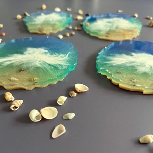 May include: Four resin coasters with a beach-themed design. Each coaster features a blue and white wave pattern over a sandy base, with small seashells embedded. The coasters are round with a scalloped edge, and are surrounded by additional seashells.