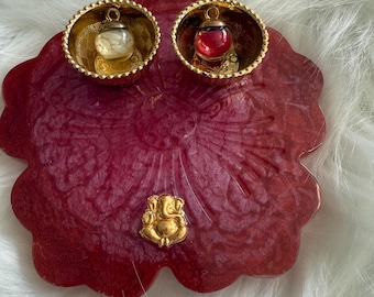 May include: A red resin tray featuring a gold Ganesh figure and two gold-rimmed bowls. Each bowl contains a small glass orb, one with a white interior and the other with a red interior. The tray rests on a white, fluffy surface.