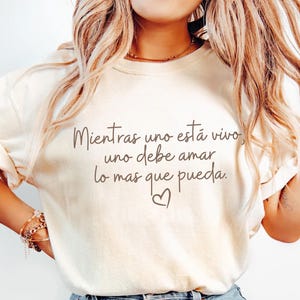 Puede incluir: Camiseta color crema con el texto en español "Mientras uno está vivo, uno debe amar lo mas que pueda" y un símbolo de corazón. La camiseta tiene mangas cortas y se combina con vaqueros azules.