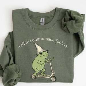 May include: Olive green sweatshirt with the text "Off to commit nana foolery." The graphic features a green frog wearing a wizard hat and riding a scooter. The sweatshirt is a Gildan brand.