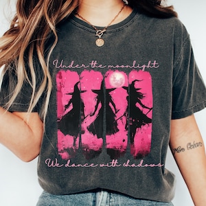 Puede incluir: Una camiseta gris carbón de manga corta con un gráfico de tres siluetas de brujas negras sobre un fondo de pinceladas rosa brillante con una luna llena. El texto Under the moonlight We dance with shadows aparece en una fuente cursiva rosa claro.