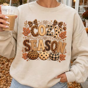 Puede incluir: Una sudadera beige de cuello redondo con un estampado gráfico que incluye el texto COZY SEASON en letras de bloque. El diseño presenta patrones de estampado de leopardo, cuadros y lunares, acentuados con ilustraciones de lazos, hojas de otoño y calabazas.