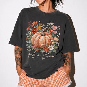 Puede incluir: Una camiseta gris carbón de manga corta con un gráfico central de una gran calabaza naranja rodeada de flores coloridas en tonos rojo, rosa, amarillo y blanco. El texto "Fall in Bloom" está impreso en letra cursiva debajo de la calabaza.