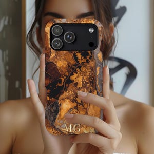 Puede incluir: Una funda de teléfono con un diseño veteado en tonos marrón, dorado y negro. La funda tiene un módulo de cámara negro con tres lentes y un flash. Las iniciales "MG" son visibles en dorado en la parte inferior.
