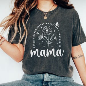 Puede incluir: Una camiseta gris oscuro con un diseño gráfico blanco que presenta una flor con un círculo de texto alrededor que dice "Amada, trabajadora, desinteresada, protectora, mamá".