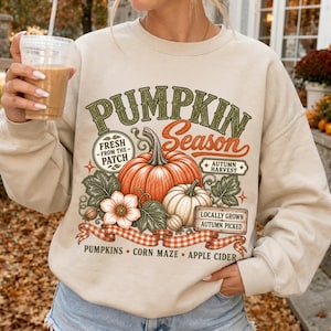 Puede incluir: Una sudadera de cuello redondo en color beige con un estampado gráfico que incluye el texto PUMPKIN Season, Fresh from the patch, Autumn harvest y Locally grown autumn picked, junto a ilustraciones de calabazas, hojas y una cinta a cuadros.