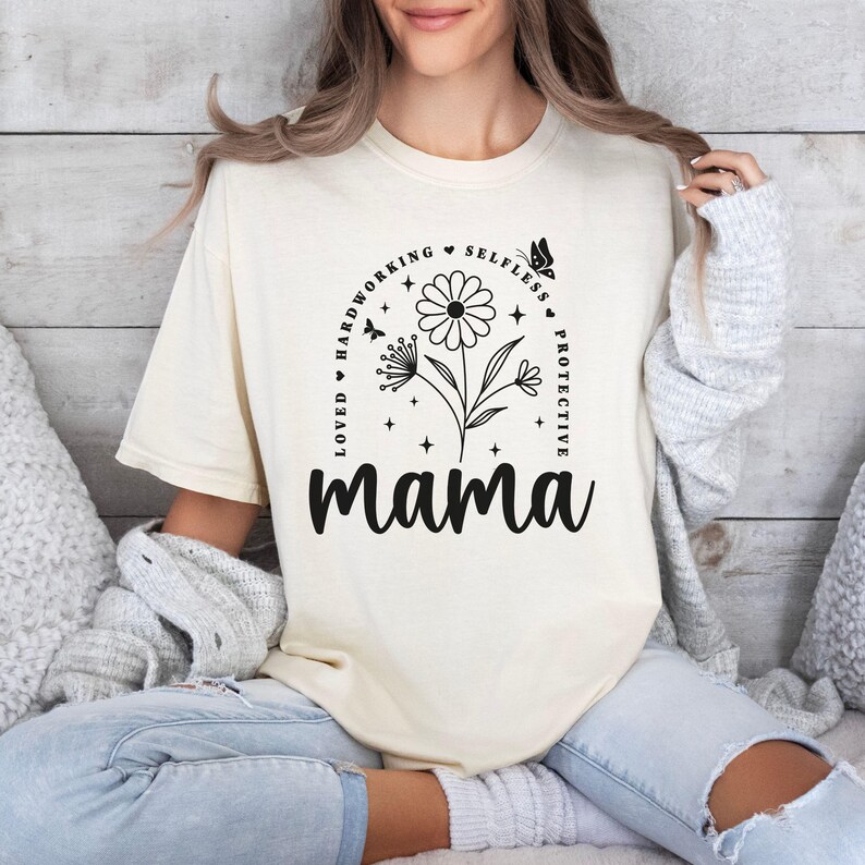 以下が含まれることがあります： 黒いグラフィックデザインが施された白いTシャツ。花が描かれており、その周りを「Loved, Hardworking, Selfless, Protective, Mama」というテキストの円が囲んでいます。