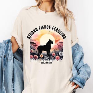 Puede incluir: Camiseta color crema con un gráfico de una silueta de león negro sobre un fondo de puesta de sol con las palabras "STRONG FIERCE FEARLESS". Acentos florales y el texto "EST. MMXXV" completan el diseño.