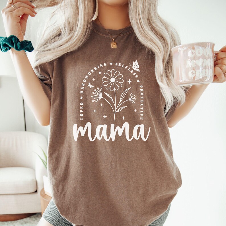 以下が含まれることがあります： 白い蝶と花柄の白いグラフィックデザインが施された茶色のTシャツ。テキストは「Loved Hardworking Selfless Protective Mama」です。