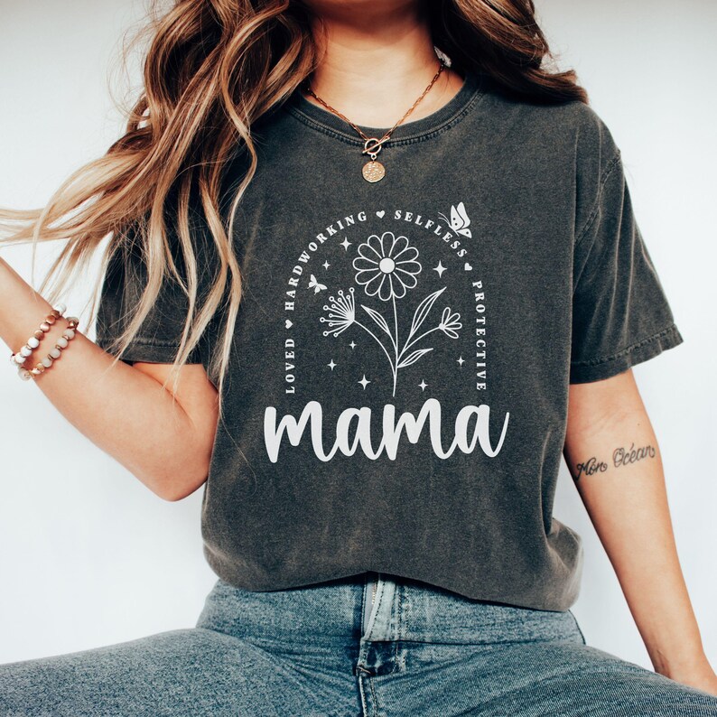 Puede incluir: Una camiseta gris oscuro con un diseño gráfico blanco que presenta una flor con un círculo de texto alrededor que dice "Amada, trabajadora, desinteresada, protectora, mamá".