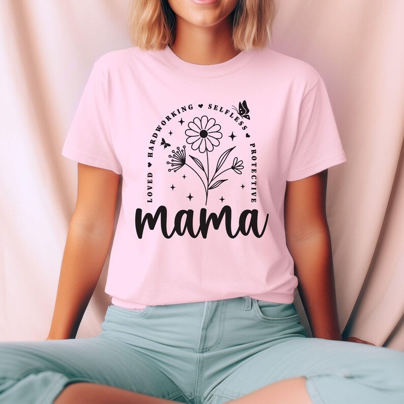 以下が含まれることがあります： 黒い花柄のデザインと「Loved, Hardworking, Selfless, Protective, Mama」というテキストが書かれたピンクのTシャツ。