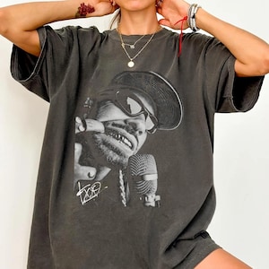 Puede incluir: Camiseta gris oscuro con un gráfico en blanco y negro de un hombre con gafas de sol y sombrero, sosteniendo un micrófono. La camiseta tiene un aspecto vintage y desgastado, con la firma del artista en blanco.