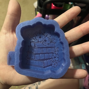 Könnte beinhalten: Eine blaue Silikonform mit einem Stapel Bücher mit floralen Akzenten. Die Form ist mit einem detaillierten Relief der Bücher und Blumen gestaltet, ideal für Bastelprojekte. Die Form wird in einer Hand gehalten.