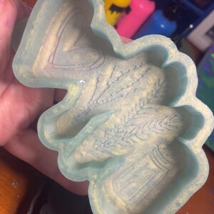 以下が含まれることがあります： ハート、小麦、塩入れのデザインが施された、水色のシリコンモールド。モールドは金色の輝きがあり、手で持たれています。黒いモールドが前景にあります。